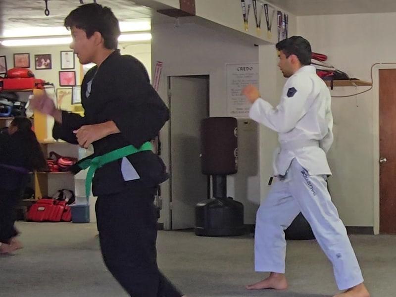 Two teens practising kicks across the floor, one in a black gi with a green belt, one in a white gi