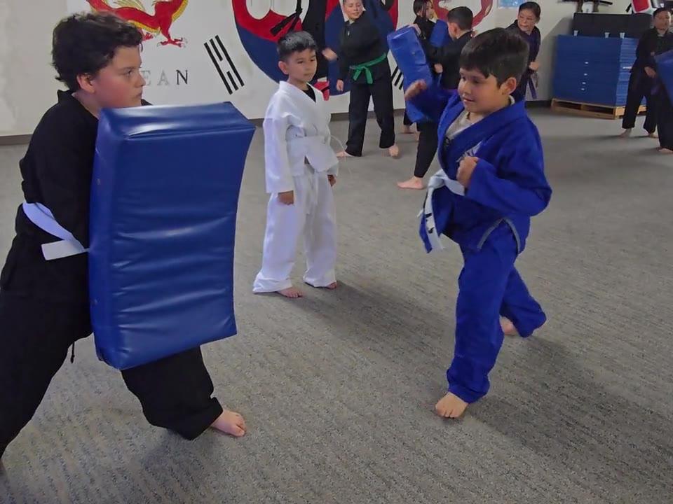 Little Tigers student in a blue gi kicking a shield held by an older classmate at the Indio dojo
