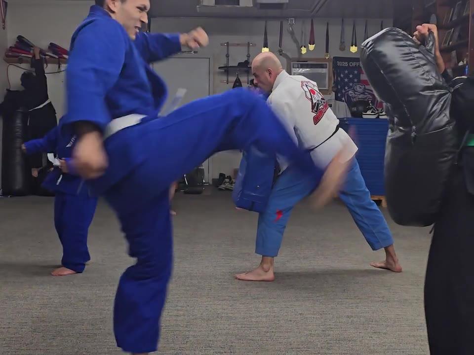Student in a blue gi throwing a kick at a shield held by a classmate, an instructor in a white gi behind