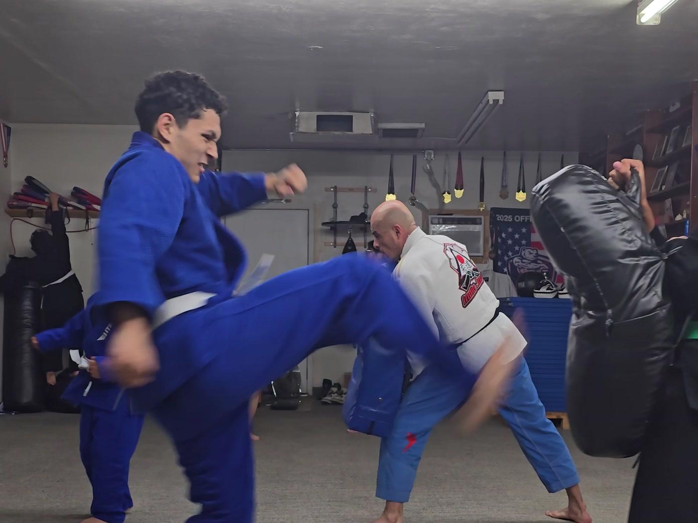 Teen in a blue gi side kicking into a body shield held by a partner